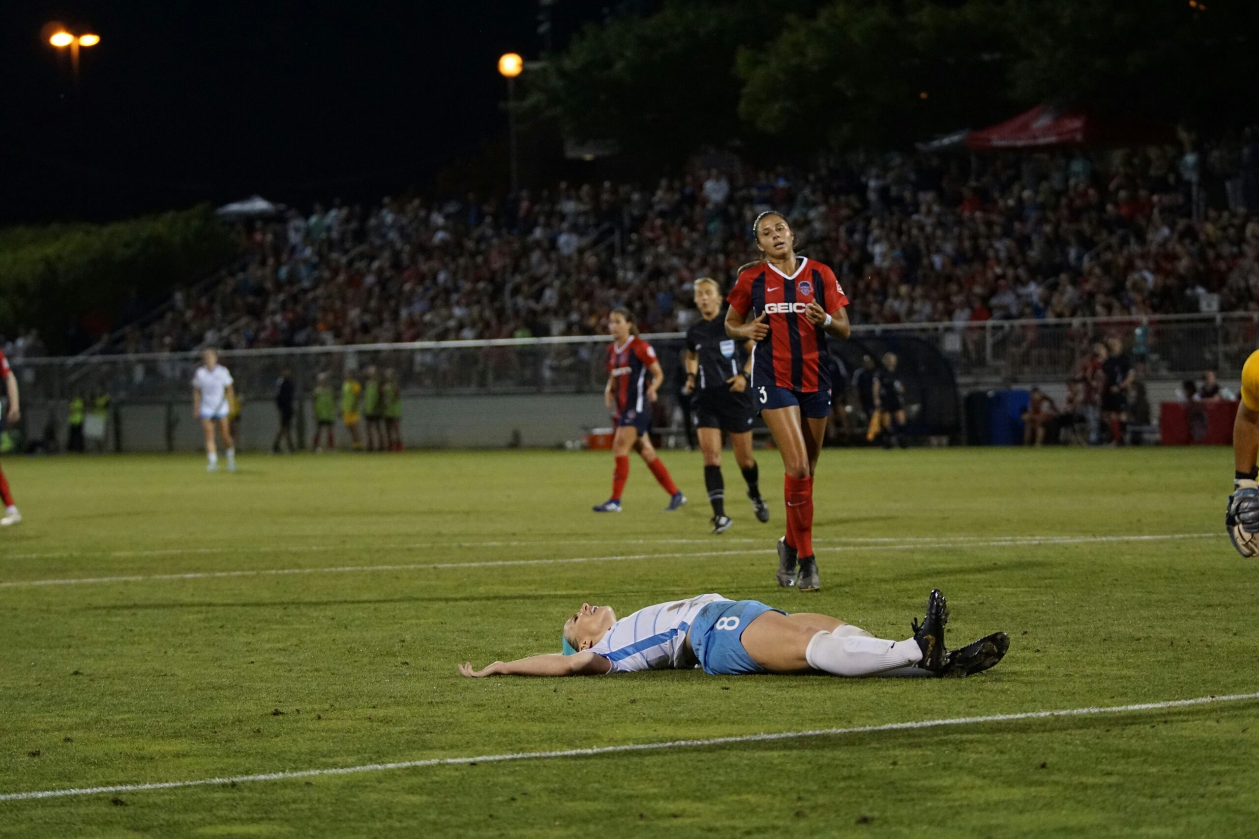 A female soccer player lying down on a green pitch alondside her running opponent: stressors to avoid on a game day