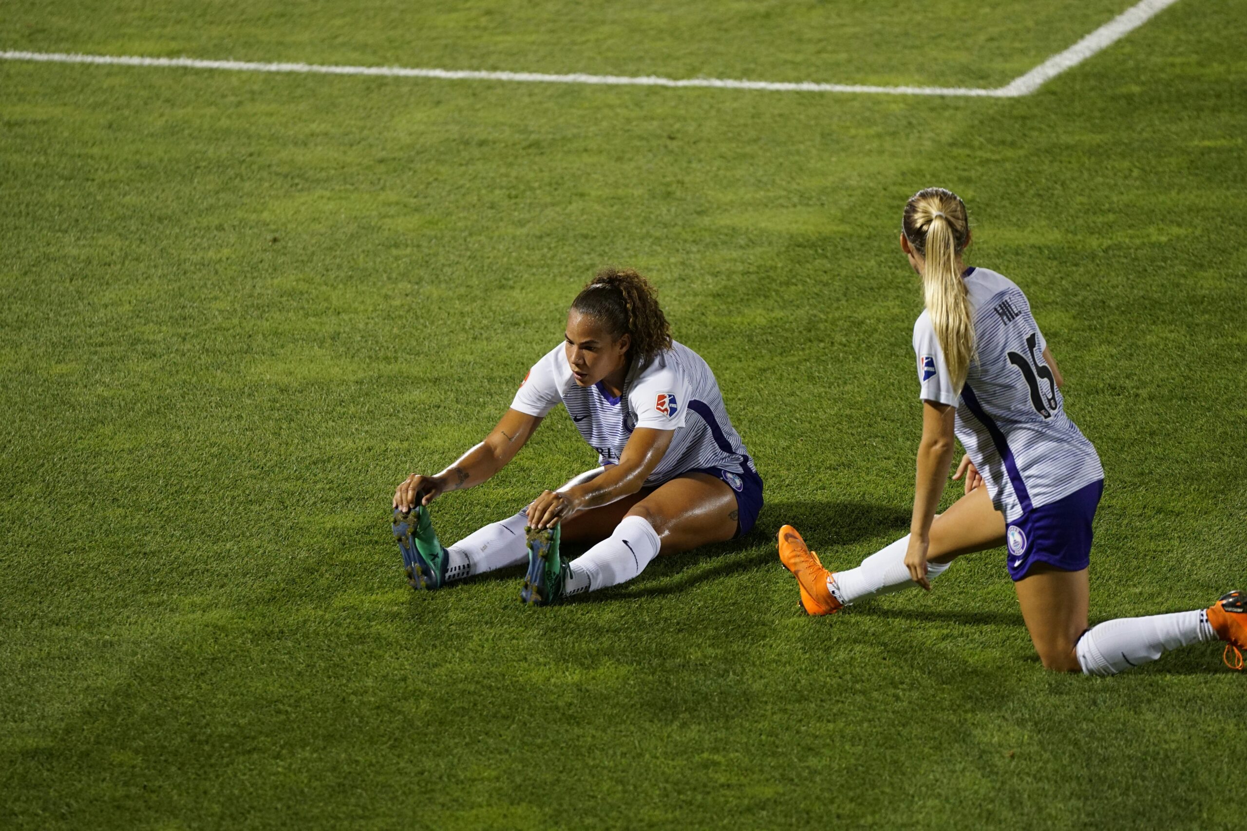 Two women soccer players stretching legs on a green lawn: Cardio workouts