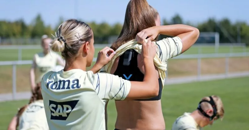 two soccer babes one adjust a GPS vest for her team mate on a green soccer pitch: kinexon-sports GPS Vests