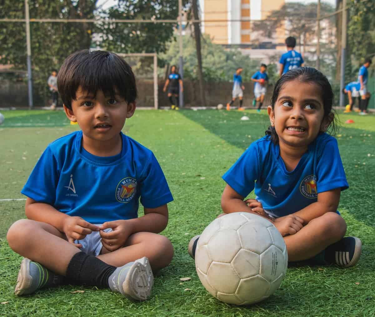 A soccer young girl alongside a boy both in polo shrt sitting near a soccer ball on a green pitch during daytime: best age for young girls to begin soccer