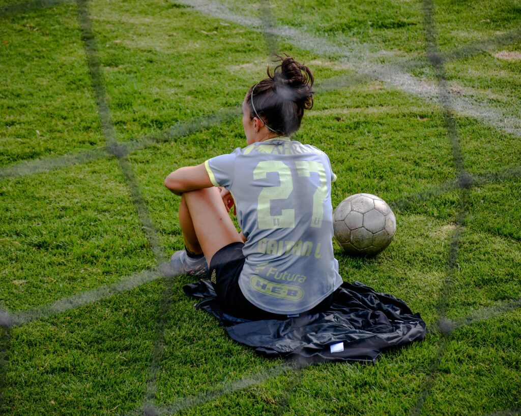 A female soccer goalkeeper siting on a green pitch alongside a soccer ball during daytime: How to Balance Soccer with School