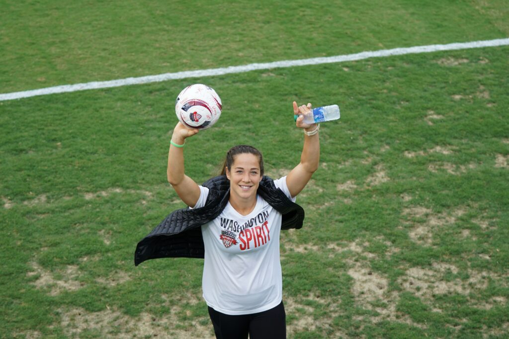 A soccer woman displaying a soccer ball and water bottle wearing a Washington Spirits Tshirt on a green soccer pitch during daytime: Women's Football Career Guide