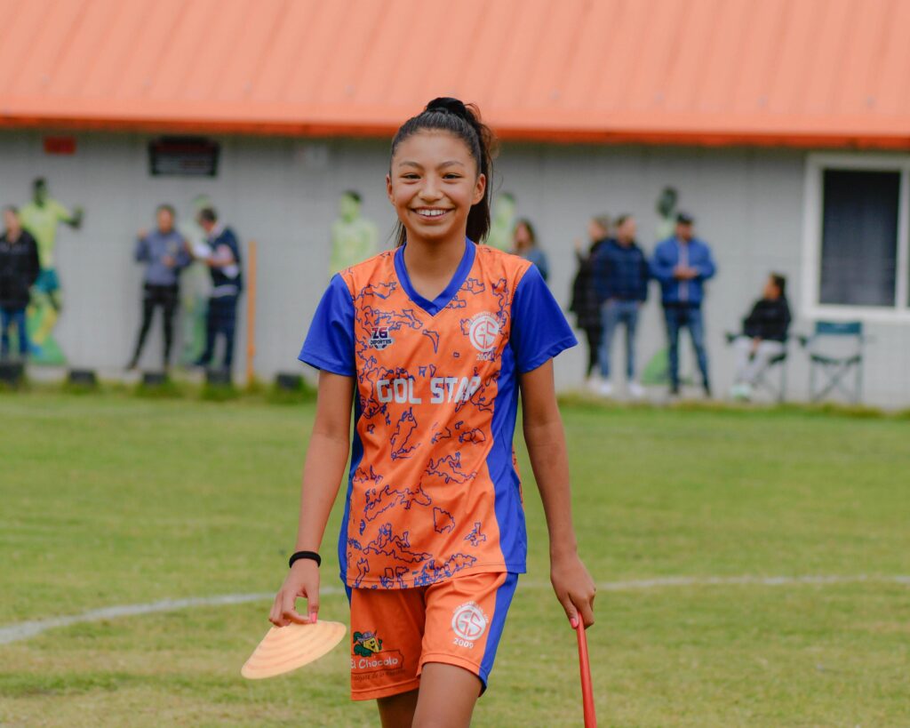 A young female soccer player smiling carrying a corn on a green pitch during day time: How to Balance Soccer with School