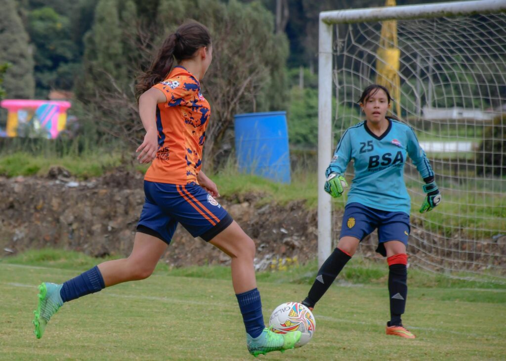 A soccer girl striker near an oppents feamle goalkeepr standing near a goal post